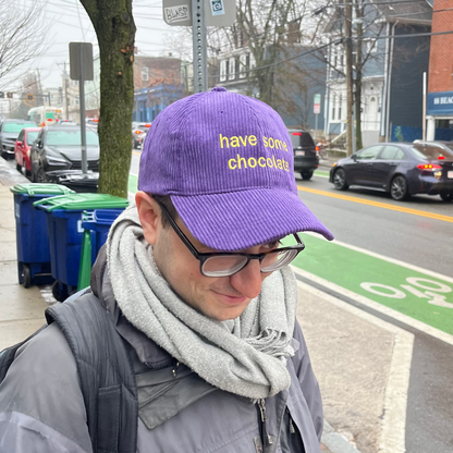 hat on head in city, reads 'have some chocolate' in yellow on purple corduroy hat